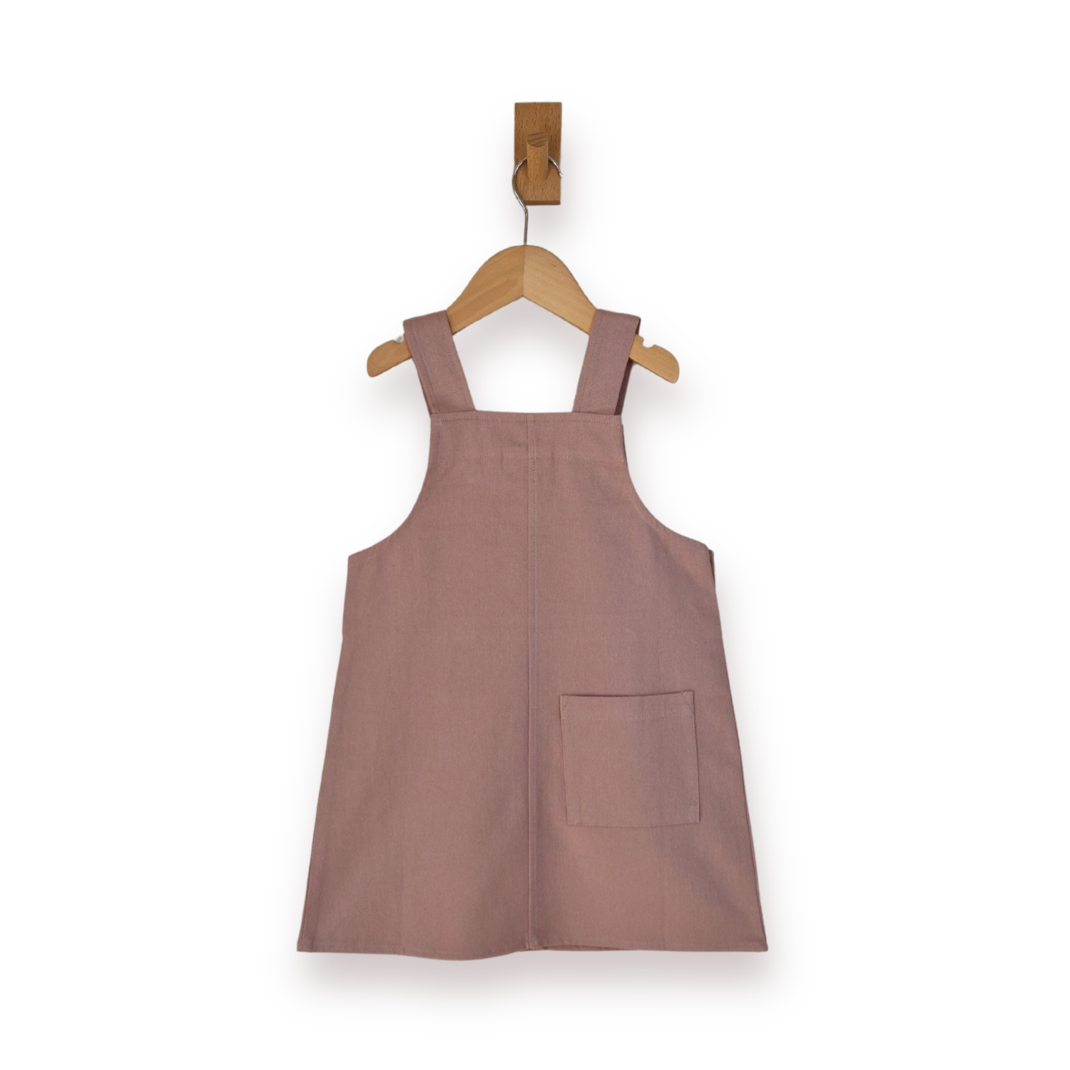 A dusty rose pink dungaree dress with a single back patch pocket, hanging on a wooden hanger against a plain white background.
