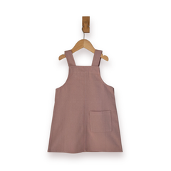 A dusty rose pink dungaree dress with a single back patch pocket, hanging on a wooden hanger against a plain white background.