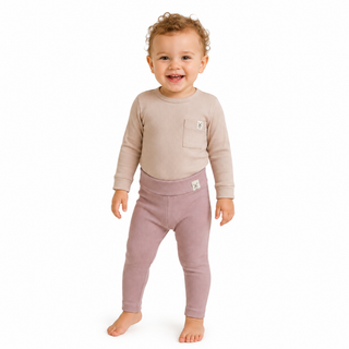 A smiling toddler girl standing barefoot against a plain white background, wearing a light beige long-sleeve top with a chest pocket and dusty pink leggings.