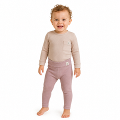 A smiling toddler girl standing barefoot against a plain white background, wearing a light beige long-sleeve top with a chest pocket and dusty pink leggings.
