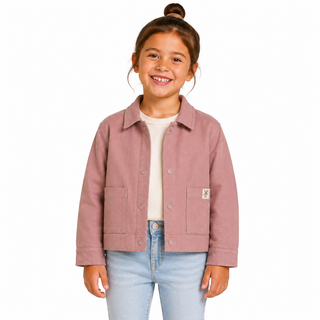 A young girl with her hair in a bun is smiling and wearing a pink jacket over a cream t-shirt and light blue jeans, photographed against a solid white background.