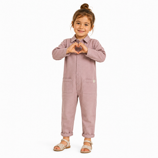 A young child wearing a dusty one-piece jumpsuit with a button-up front, collar, and front pockets, standing against a plain white background and forming a heart shape with their hands.