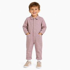 A young boy smiling and wearing a dusty rose pink button-up boilersuit with front pockets, styled with beige sneakers, standing against a plain white background.