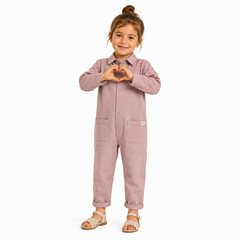 A young child wearing a dusty one-piece jumpsuit with a button-up front, collar, and front pockets, standing against a plain white background and forming a heart shape with their hands.
