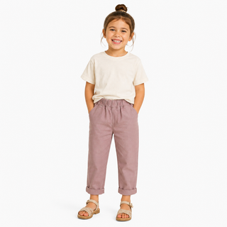 A young girl with her hair in a bun stands against a light gray background, wearing a cream t-shirt, pink cropped chinos, and beige sandals.