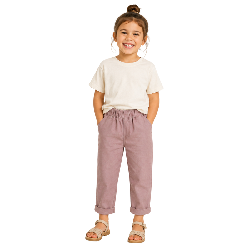 A young girl with her hair in a bun stands against a light gray background, wearing a cream t-shirt, pink cropped chinos, and beige sandals.
