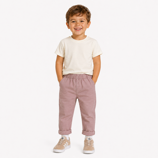 A young boy smiling and standing against a light studio background, wearing a cream short-sleeve t-shirt, dusty rose elastic-waist pants, and Adidas trainers.