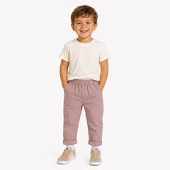 A young boy smiling and standing against a light studio background, wearing a cream short-sleeve t-shirt, dusty rose elastic-waist pants, and Adidas trainers.