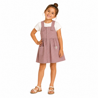 Girl wearing pink dungaree pinafore style dress over a white short sleeve tee, with beige sandals, against a white background. 