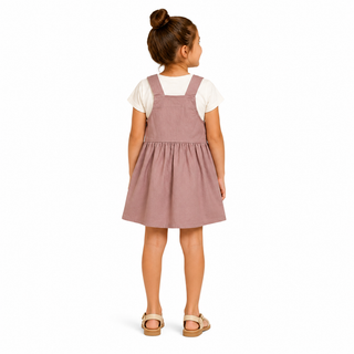 A young girl with her hair in a bun is shown from the back, wearing a dusty rose pink corduroy pinafore dress over a white t-shirt and beige sandals, set against a plain white background.