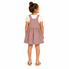 A young girl with her hair in a bun is shown from the back, wearing a dusty rose pink corduroy pinafore dress over a white t-shirt and beige sandals, set against a plain white background.