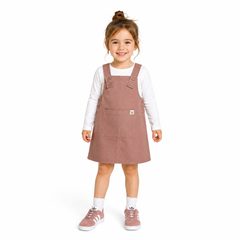A young girl with her hair in a bun stands against a white background wearing a dusty rose corduroy pinafore dress over a white long-sleeve shirt, white socks, and pink trainers.