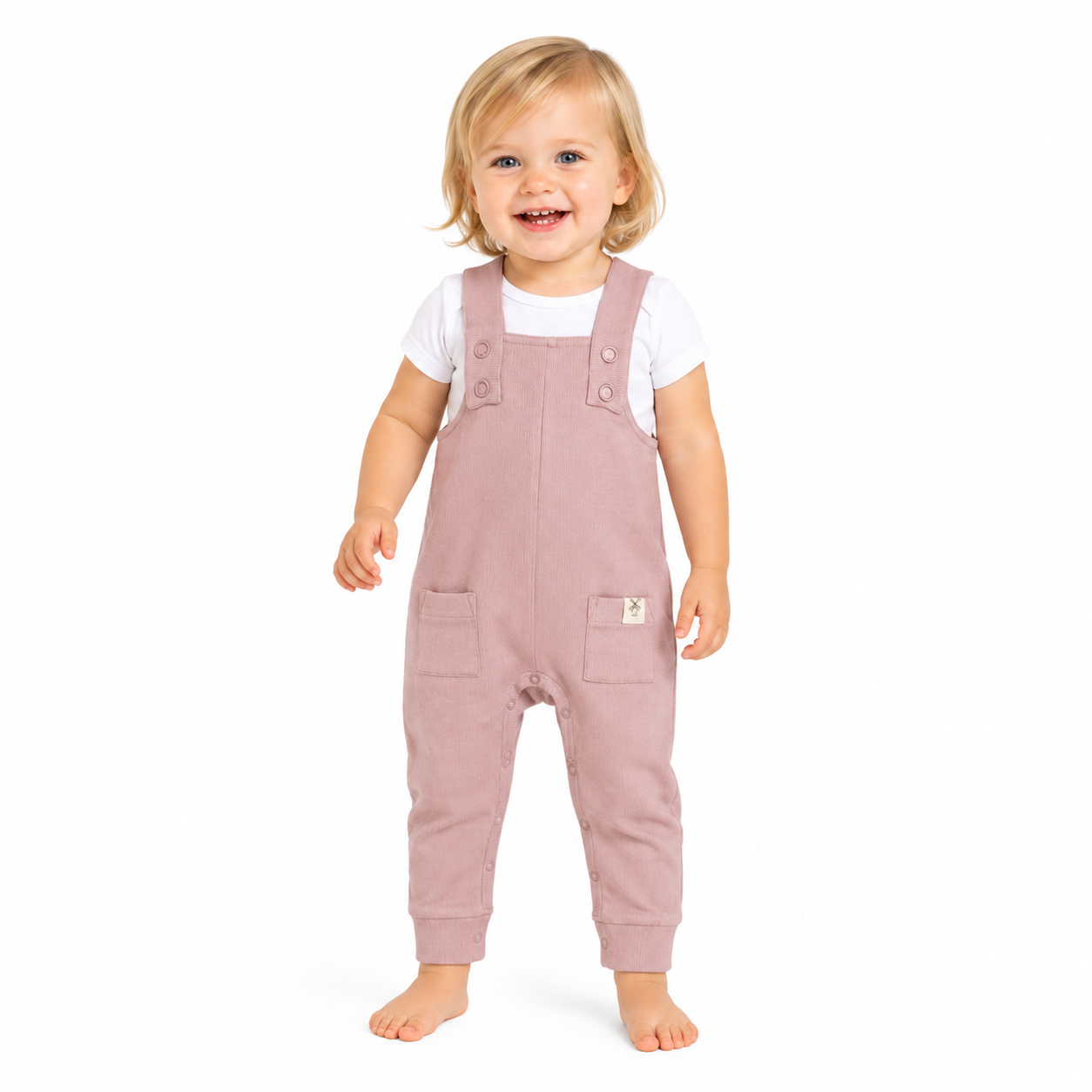A smiling toddler standing barefoot on a white background, wearing dusty pink dungarees over a white short-sleeve shirt.