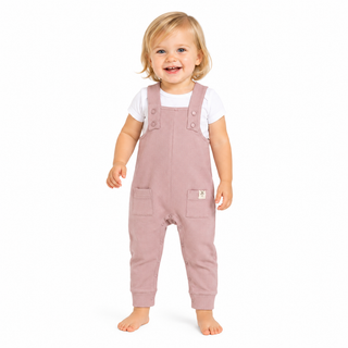 A smiling toddler standing barefoot on a white background, wearing dusty pink dungarees over a white short-sleeve shirt.
