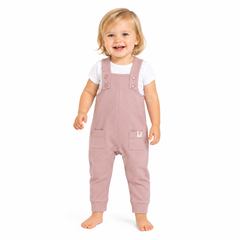 A smiling toddler standing barefoot on a white background, wearing dusty pink dungarees over a white short-sleeve shirt.
