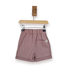 A pair of dusty rose shorts with a gathered elastic waistband, rolled hems, and a side pocket, hanging from a wooden hanger on a white background.