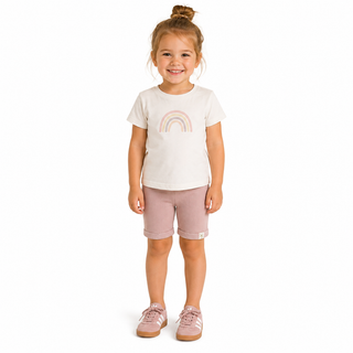 A young girl with her hair in a bun stands against a white background wearing a white t-shirt with a pink rainbow graphic, dusty pink shorts, and matching pink trainers.