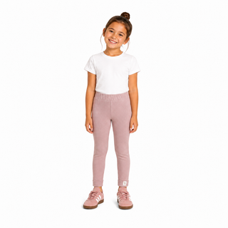 A young girl with her hair in a bun stands against a solid white background, wearing dusty pink leggings, a white t-shirt, and matching pink Adidas Gazelles.