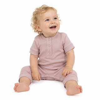 A smiling baby with curly blonde hair sits on a white background wearing a pink short-sleeve romper with a buttoned neckline.