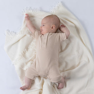 Small baby modelling short playsuit in beige whilst laying on a cream blanket