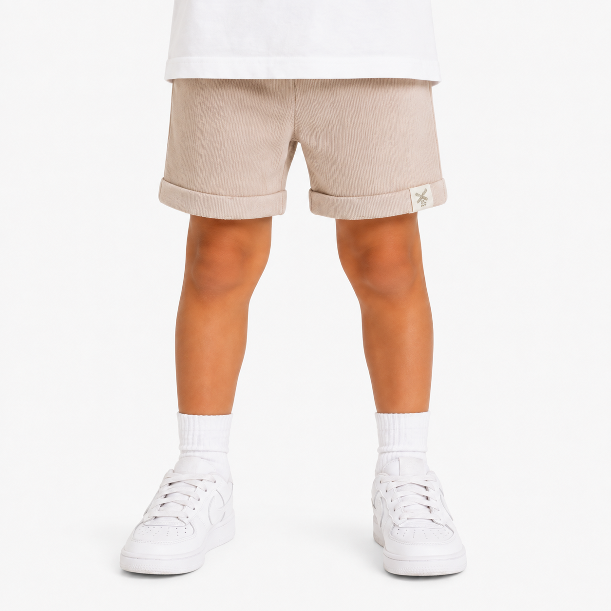A child's legs wearing light beige shorts with rolled cuffs, white socks, and white trainers, shown from the waist down on a white studio background.