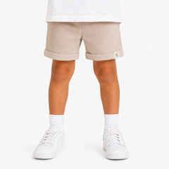 A child's legs wearing light beige shorts with rolled cuffs, white socks, and white trainers, shown from the waist down on a white studio background.