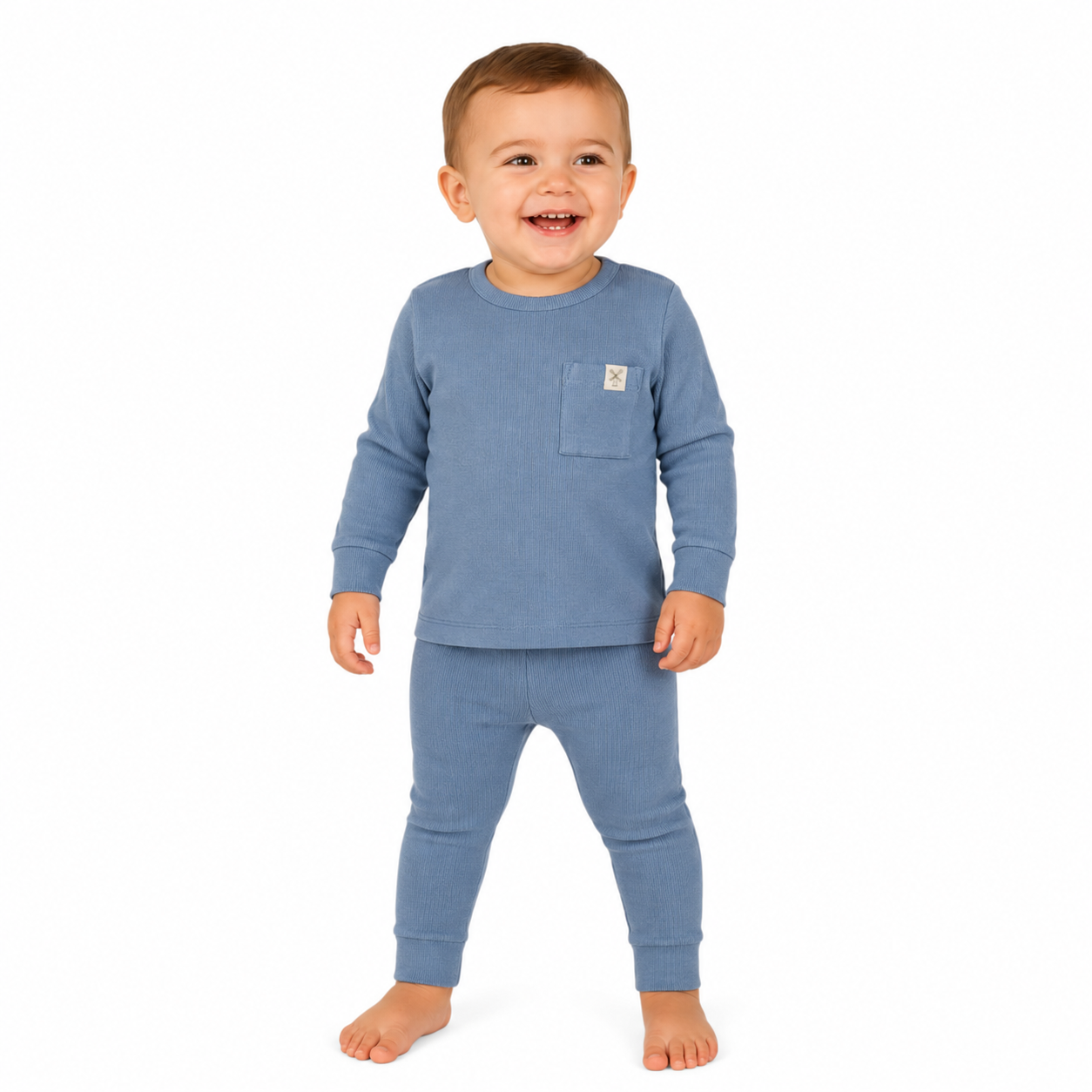 A smiling toddler standing barefoot against a plain white background, wearing a blue long-sleeve top with a chest pocket and matching blue leggings. A small square brand patch is visible on the pocket.