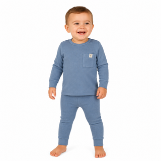 A smiling toddler standing barefoot against a plain white background, wearing a blue long-sleeve top with a chest pocket and matching blue leggings. A small square brand patch is visible on the pocket.