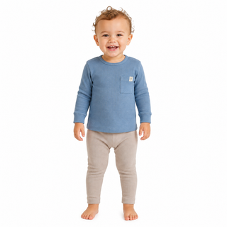 A smiling toddler standing barefoot against a white background, wearing a blue long-sleeve ribbed top with a chest pocket and light beige leggings.