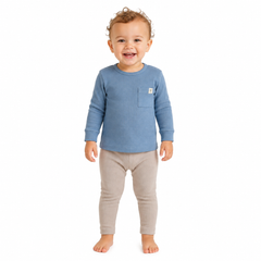 A smiling toddler standing barefoot against a white background, wearing a blue long-sleeve ribbed top with a chest pocket and light beige leggings.