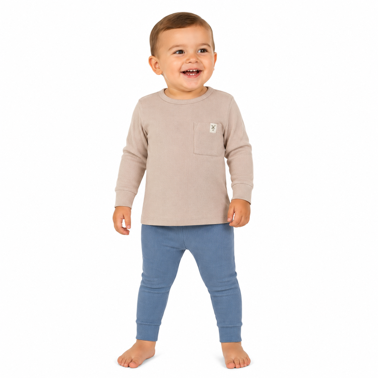 A young boy standing against a plain white background, wearing a light beige long-sleeve top with a chest pocket and blue leggings.