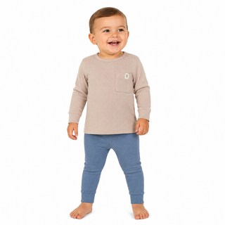 A young boy standing against a plain white background, wearing a light beige long-sleeve top with a chest pocket and blue leggings.
