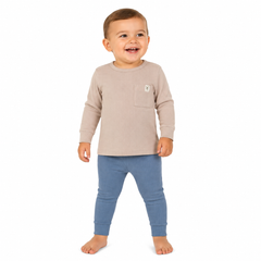A young boy standing against a plain white background, wearing a light beige long-sleeve top with a chest pocket and blue leggings.