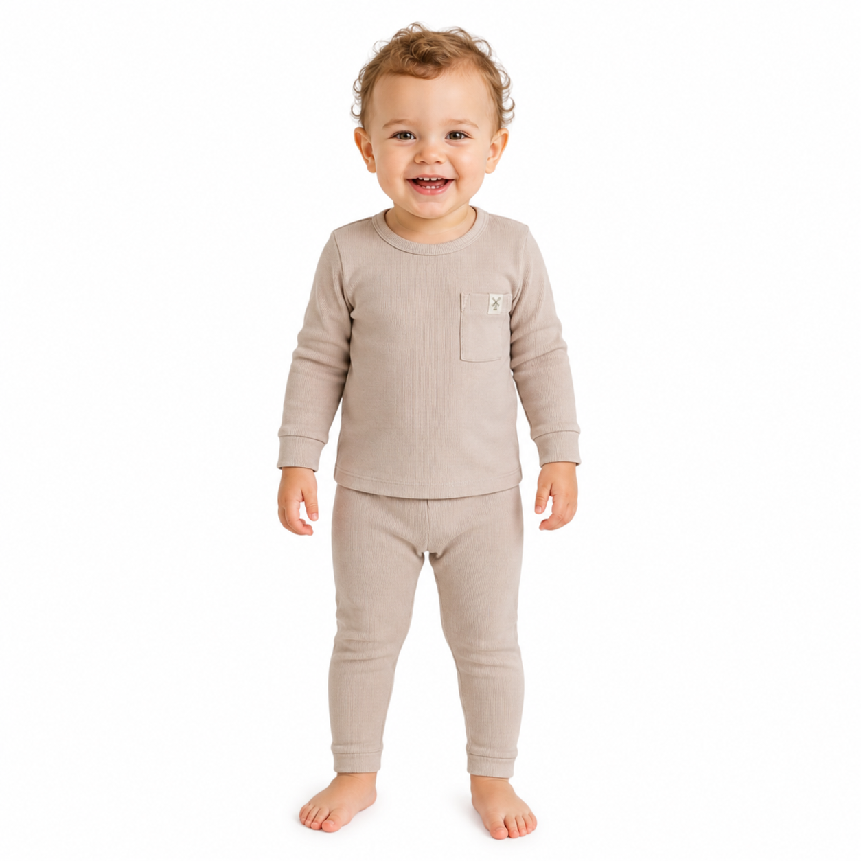 A smiling toddler girl standing barefoot against a plain white background, wearing a beige long-sleeve ribbed top with matching leggings and a small chest pocket.