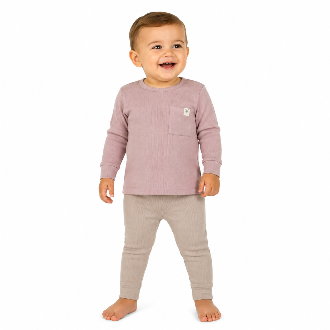 A smiling toddler standing barefoot against a white background, wearing a pink long-sleeve top with a chest pocket and light beige pants.