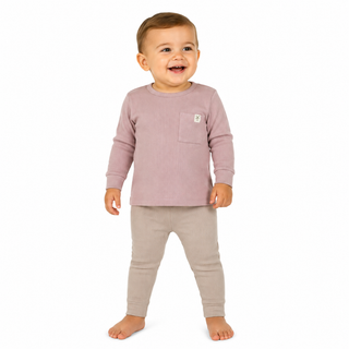 A smiling toddler standing barefoot against a white background, wearing a pink long-sleeve top with a chest pocket and light beige pants.