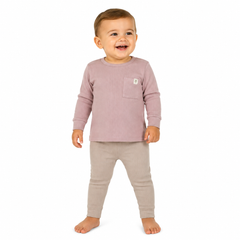 A smiling toddler standing barefoot against a white background, wearing a pink long-sleeve top with a chest pocket and light beige pants.