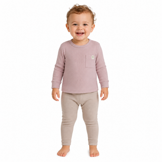 A smiling toddler standing barefoot against a plain white background, wearing a dusty pink long-sleeve top with a chest pocket and light beige pants.