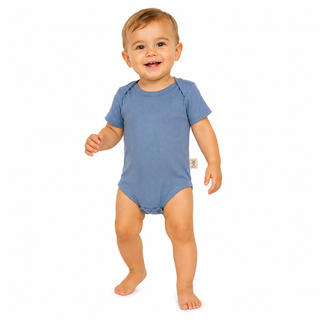 A smiling toddler standing barefoot on a white background, wearing a short-sleeve blue bodysuit with a small beige Aneby windmill tag on the side.