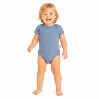 A smiling girl standing barefoot against a plain white background, wearing a short-sleeve blue bodysuit.
