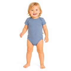 A smiling girl standing barefoot against a plain white background, wearing a short-sleeve blue bodysuit.