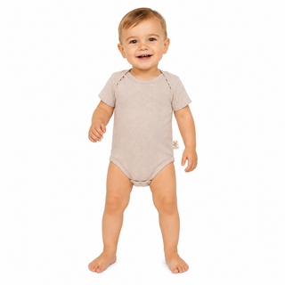 A smiling baby standing barefoot on a white background, wearing a short-sleeve, one-piece baby bodysuit in a light taupe-beige color.