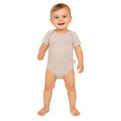 A smiling baby standing barefoot on a white background, wearing a short-sleeve, one-piece baby bodysuit in a light taupe-beige color.