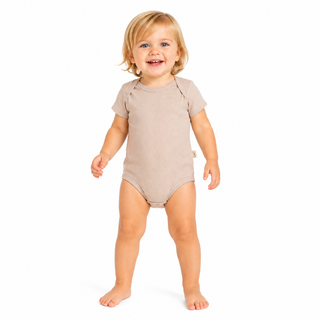 A smiling young girl standing barefoot on a white background, wearing a short-sleeve, one-piece baby bodysuit in a light beige colour.