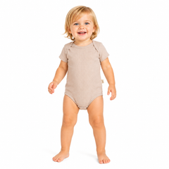 A smiling young girl standing barefoot on a white background, wearing a short-sleeve, one-piece baby bodysuit in a light beige colour.