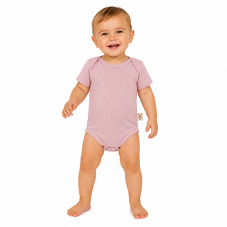 A smiling baby standing barefoot on a white background, wearing a short-sleeve dusty pink bodysuit with a small beige tag on the side.