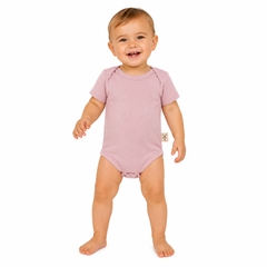 A smiling baby standing barefoot on a white background, wearing a short-sleeve dusty pink bodysuit with a small beige tag on the side.