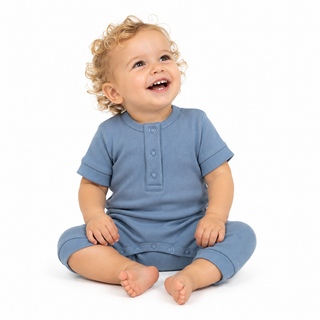 A smiling toddler with curly blonde hair sits cross-legged on a white background, wearing a blue short-sleeve romper with a buttoned placket neckline.