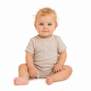 A baby with blonde hair sitting cross-legged on a white background, wearing a light beige short-sleeve romper.