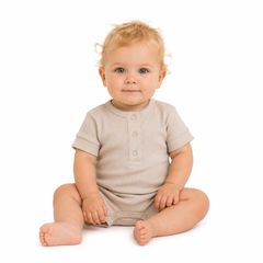 A baby with blonde hair sitting cross-legged on a white background, wearing a light beige short-sleeve romper.